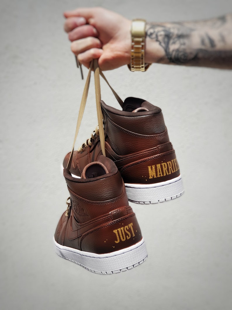 May include: A pair of brown high-top sneakers with white soles. The words "JUST" and "MARRIED" are printed in gold on the heels. The shoes are held by their laces.