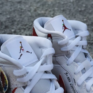 May include: White leather high-top sneakers with red and white paint splatters. The sneakers feature a white leather tongue with a black and red silhouette of a basketball player. The right shoe has the text "5 x NBA Champion" written on the side.