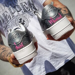 May include: Pair of custom gray and white AF-1 sneakers. The heels are inscribed with the words "I just" and "Married" in pink script. The shoes have a white sole and gray accents.