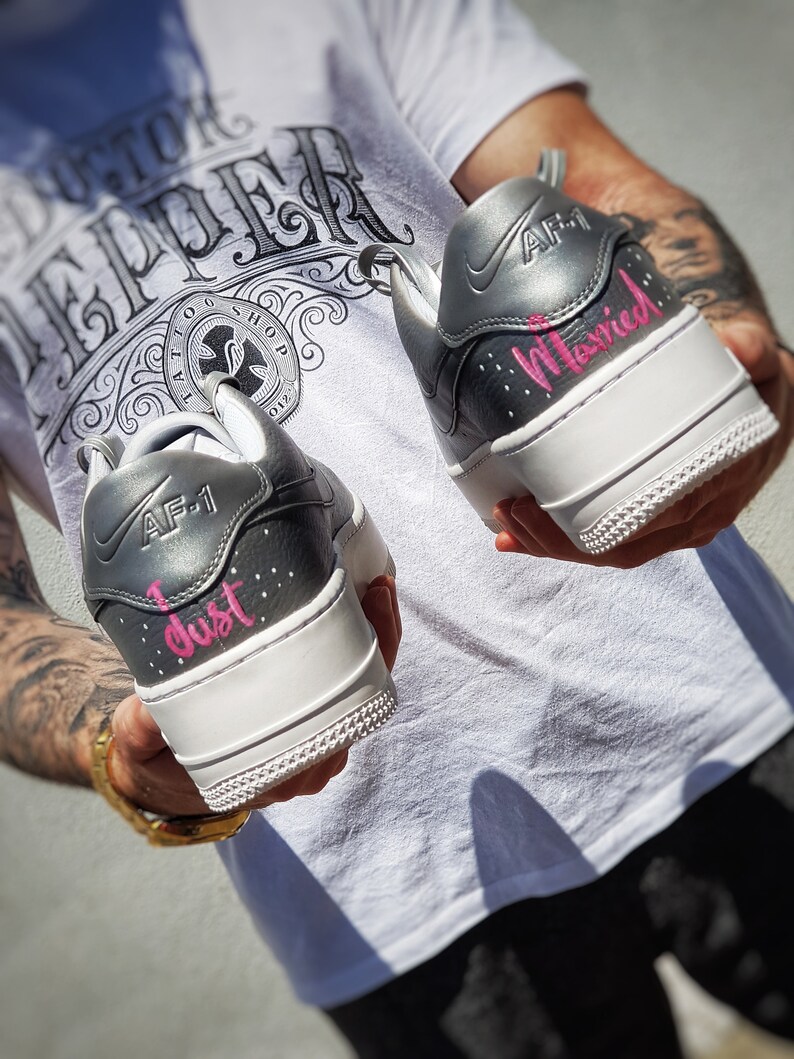 May include: Pair of custom gray and white AF-1 sneakers. The heels are inscribed with the words "I just" and "Married" in pink script. The shoes have a white sole and gray accents.