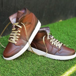 May include: A pair of brown leather high-top sneakers with white soles and tan laces. The sneakers have a classic design with the Nike swoosh logo on the side. The shoes are displayed on a bed of green grass.