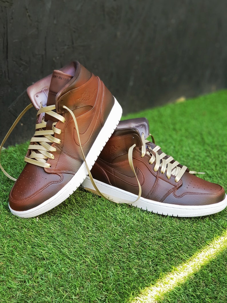 May include: A pair of brown leather high-top sneakers with white soles and tan laces. The sneakers have a classic design with the Nike swoosh logo on the side. The shoes are displayed on a bed of green grass.
