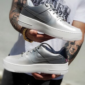 May include: Silver and white platform sneakers with the Nike swoosh logo. The shoes have a textured silver upper and a thick white sole. The laces are silver and the design is modern and stylish.