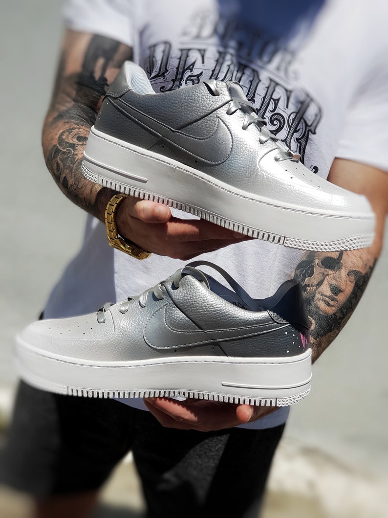 May include: Silver and white platform sneakers with the Nike swoosh logo. The shoes have a textured silver upper and a thick white sole. The laces are silver and the design is modern and stylish.