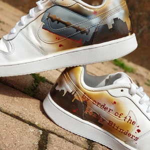 May include: White leather trainers with a custom paint design featuring a razor blade, a cityscape, and the text "By order of the Peaky Blinders."