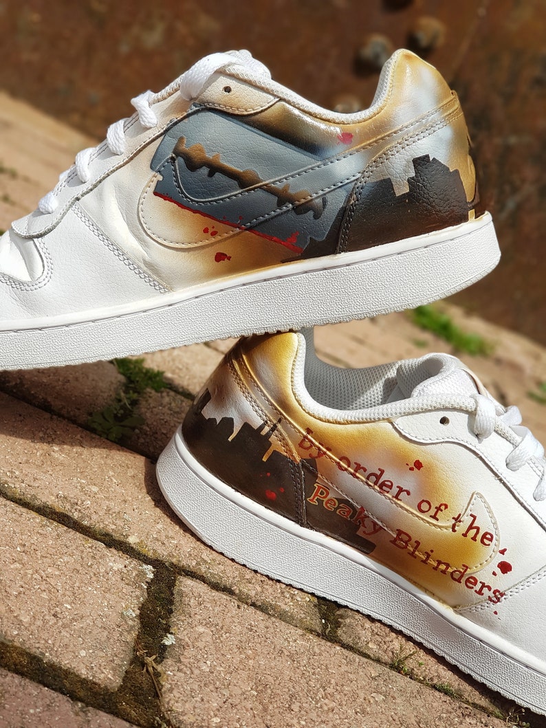May include: White leather trainers with a custom paint design featuring a razor blade, a cityscape, and the text "By order of the Peaky Blinders."