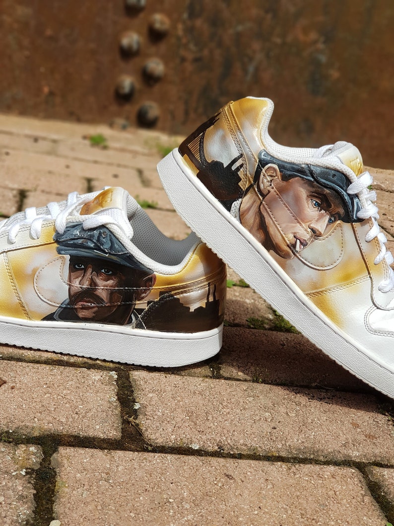 May include: A pair of white trainers with a custom design featuring a portrait of a man wearing a cap and a city skyline in the background. The trainers have a yellowed effect and a white swoosh logo.