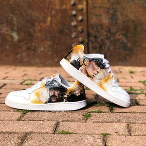 May include: A pair of white trainers with a custom painted design. The design features a portrait of a man wearing a hat and a cityscape in the background. The trainers are on a brick surface.