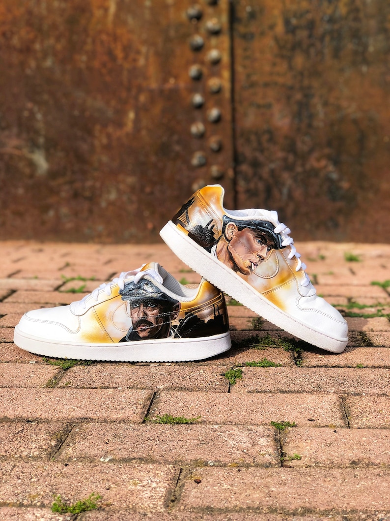 May include: A pair of white trainers with a custom painted design. The design features a portrait of a man wearing a hat and a cityscape in the background. The trainers are on a brick surface.