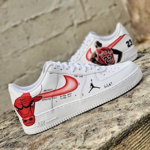 May include: White sneakers with custom artwork. One shoe features a red bull illustration and text including "MICHAEL" and "PAPADEMETRIOU". The other shoe displays a basketball player in a red jersey with the number "23". The shoes also have the text "GOAT".