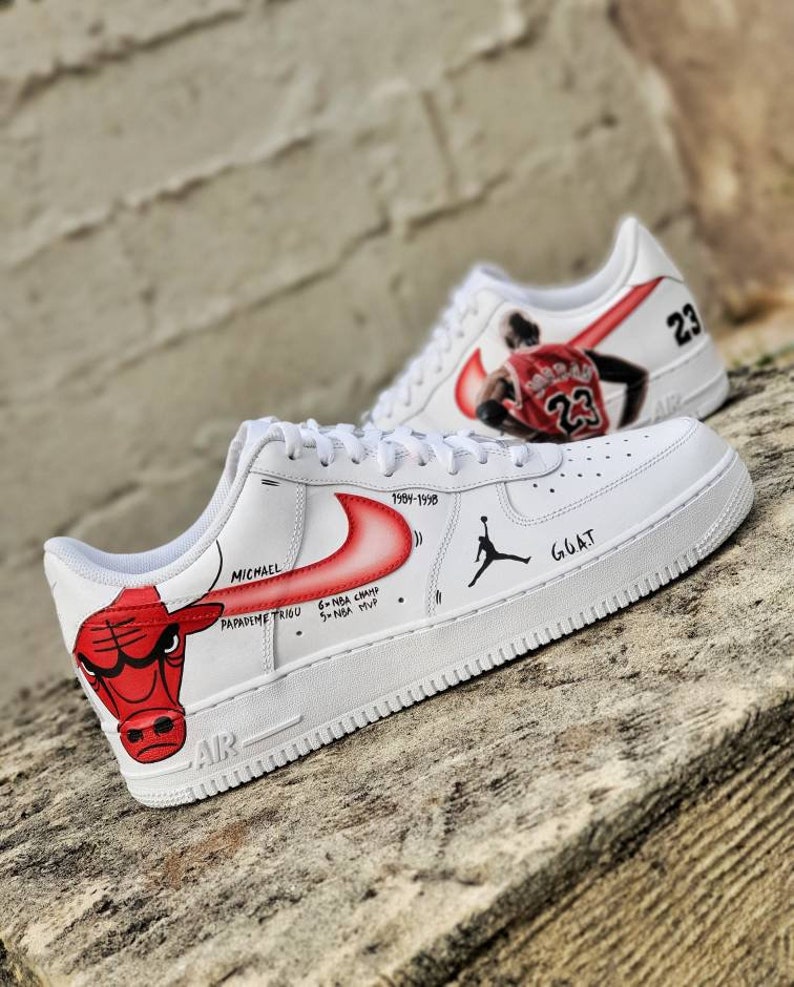 May include: White sneakers with custom artwork. One shoe features a red bull illustration and text including "MICHAEL" and "PAPADEMETRIOU". The other shoe displays a basketball player in a red jersey with the number "23". The shoes also have the text "GOAT".