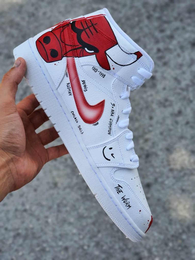 May include: White high-top sneakers with a red and black cartoon bull on the side. The text "Dennis Rodman" and "Chicago Bulls" are written on the side of the shoe. The text "5x NBA Champion" and "1995-1998" are also written on the side of the shoe. The text "The Worm" is written on the bottom of the shoe.