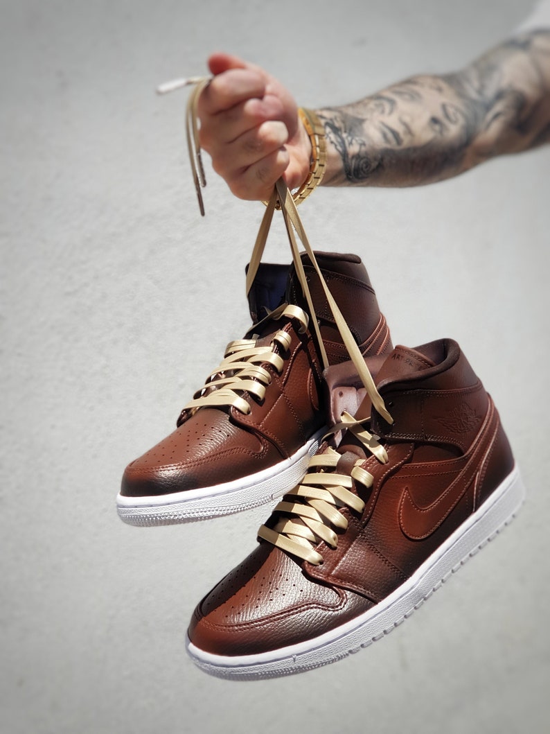 May include: A pair of brown leather high-top sneakers with white soles and tan laces. The sneakers feature a perforated toe box and a brown Nike swoosh on the side. The shoes are suspended by their laces.