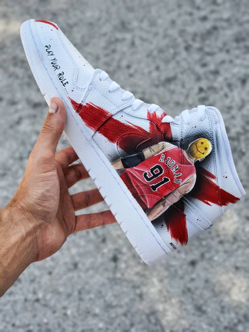May include: White high-top sneakers with red and black paint splatters. The sneakers feature a basketball player with the number 91 and the name "Rodman" on the side. The text "Play Your Role" is written on the side of the shoe.