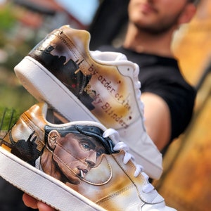 May include: White trainers with a hand-painted design featuring a man in a cap and the text "by order of the Peaky Blinders".