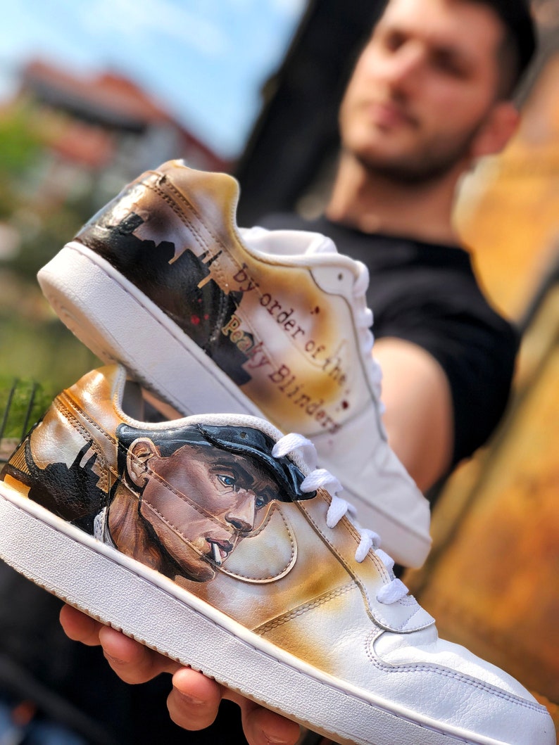 May include: White trainers with a hand-painted design featuring a man in a cap and the text "by order of the Peaky Blinders".