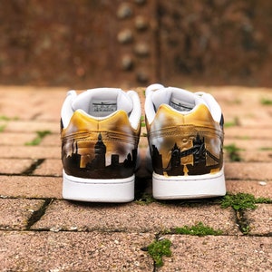 May include: A pair of white trainers with a gold paint design. The left shoe features a cityscape silhouette, while the right shoe has a silhouette of Tower Bridge in London.