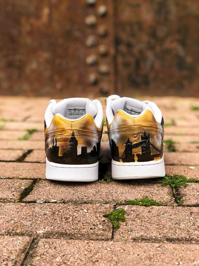 May include: A pair of white trainers with a gold paint design. The left shoe features a cityscape silhouette, while the right shoe has a silhouette of Tower Bridge in London.