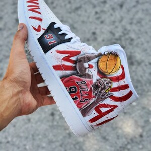 May include: White high-top sneakers with a red and black paint design featuring a basketball player in a red jersey with the number 91 and the word "BULLS". The player is holding a basketball and the words "MICHAEL" and "JORDAN" are painted on the shoe.