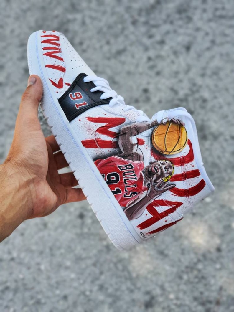 May include: White high-top sneakers with a red and black paint design featuring a basketball player in a red jersey with the number 91 and the word "BULLS". The player is holding a basketball and the words "MICHAEL" and "JORDAN" are painted on the shoe.