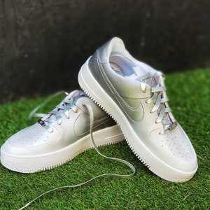 May include: Silver-colored sneakers with white soles and laces. The shoes have a textured leather upper and the Nike swoosh logo on the side. The sneakers are on a green grassy surface.
