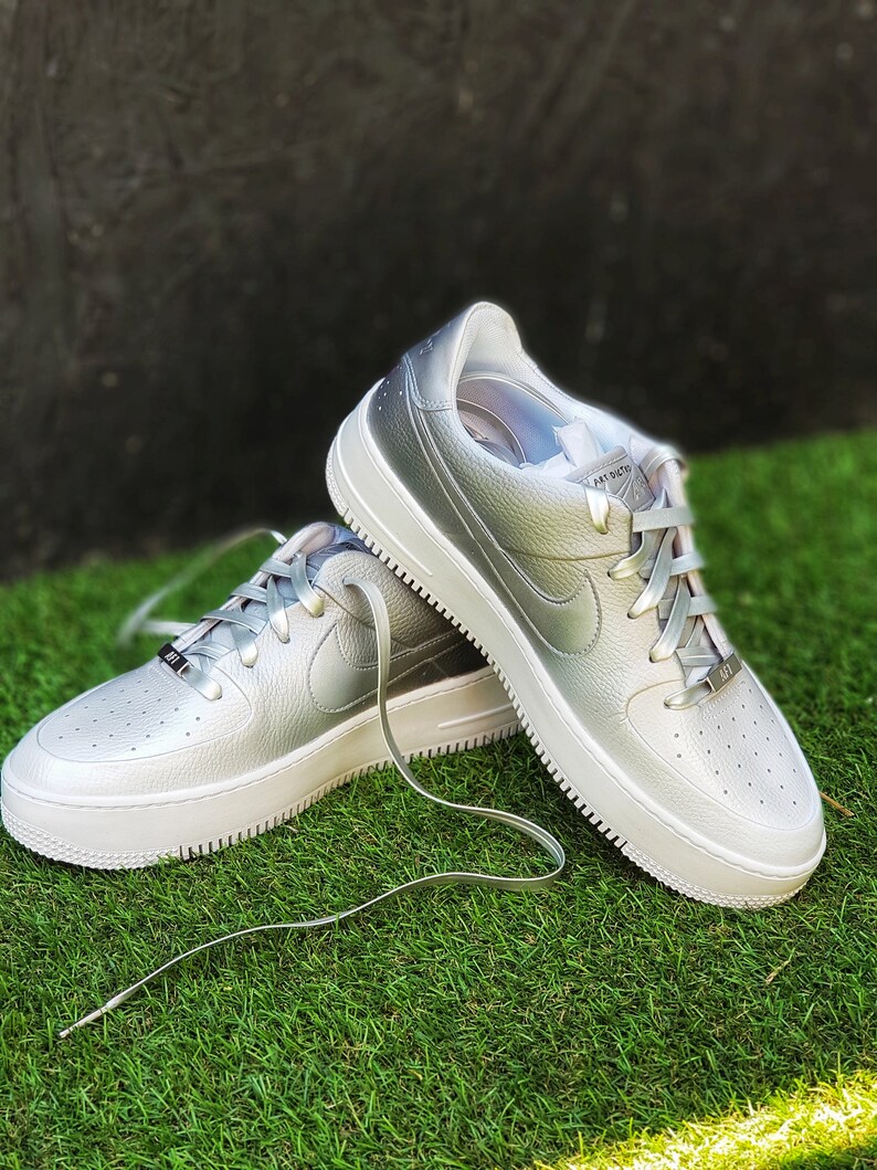 May include: Silver-colored sneakers with white soles and laces. The shoes have a textured leather upper and the Nike swoosh logo on the side. The sneakers are on a green grassy surface.