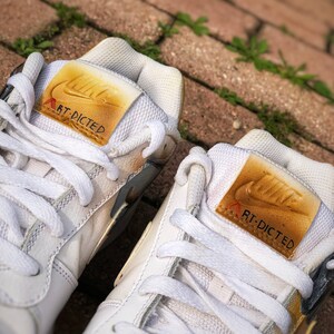 May include: A pair of white leather trainers with gold accents. The trainers have a Nike swoosh logo on the side and the text "RT-DICTED" on the tongue.