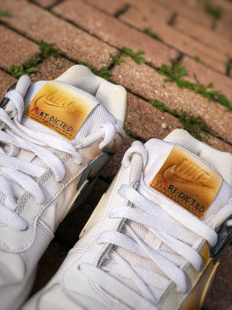 May include: A pair of white leather trainers with gold accents. The trainers have a Nike swoosh logo on the side and the text "RT-DICTED" on the tongue.