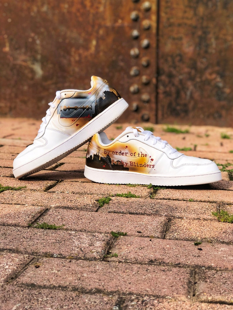 May include: A pair of white trainers with a custom design featuring a razor blade and the text "By order of the Peaky Blinders".