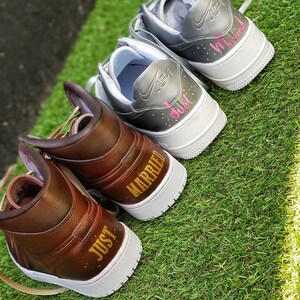 May include: Two pairs of custom sneakers, one brown and one gray, with white soles, sit on green grass. The brown sneakers say "JUST MARRIED" in gold lettering. The gray sneakers say "Just Married" in pink lettering.
