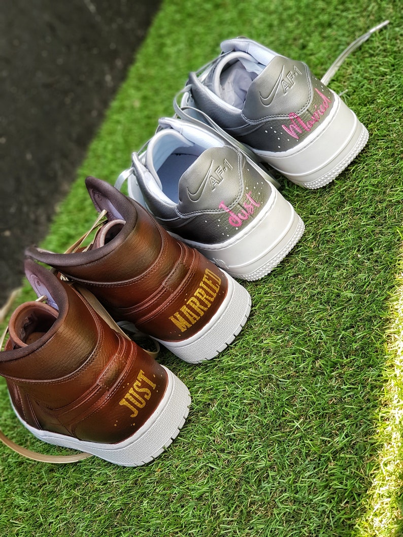 May include: Two pairs of custom sneakers, one brown and one gray, with white soles, sit on green grass. The brown sneakers say "JUST MARRIED" in gold lettering. The gray sneakers say "Just Married" in pink lettering.