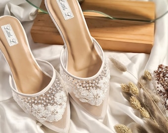 May include: A pair of white bridal heels with delicate lace and rhinestone embellishments. The heels feature a pointed toe and a block heel. The shoes are labeled "Little L".