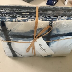 May include: A stack of fabric squares wrapped in clear plastic and tied with a raffia bow. The fabric features a watercolor-like design in shades of blue and white. A tag with the text "Boundless Fabrics" is attached.