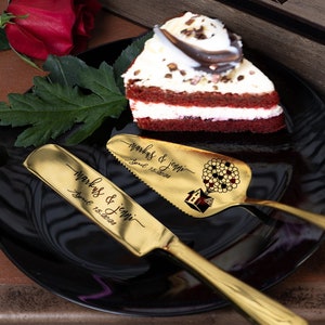 以下が含まれることがあります： 家と熱気球が彫刻された木製の箱。箱が開いており、2本の金色のケーキサーバーが置かれており、サーバーには「Mark & Jenni」と「June 12, 2023」の文字が刻まれています。