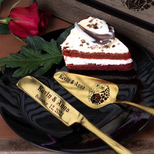 May include: A wooden box engraved with "Kevin & Anna" and a design of dice, along with two gold cake servers engraved with "Kevin & Anna" and a date. The servers are on a black plate with a slice of red velvet cake.