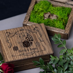 May include: A wooden ring box with a hinged lid. The lid is engraved with a design of dice and the text "Kevin & Anna 11.28.2023". The box is lined with green moss and two gold rings are inside.