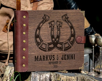 Peut inclure: Un livre d'or en bois avec une couverture en cuir marron et des accents en laiton. La couverture présente deux fers à cheval entrelacés avec un motif de nœud celtique. Le texte "MARKUS & JENNI SEPTEMBRE 20, 2022" est gravé sous les fers à cheval.