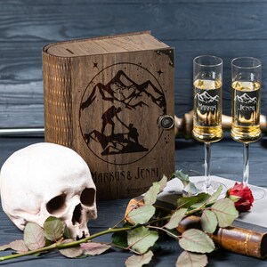 May include: A wooden box with a laser engraved design of two figures hiking in mountains. The box is closed and has a metal latch. The box is sitting on a black surface with a skull and a rose.