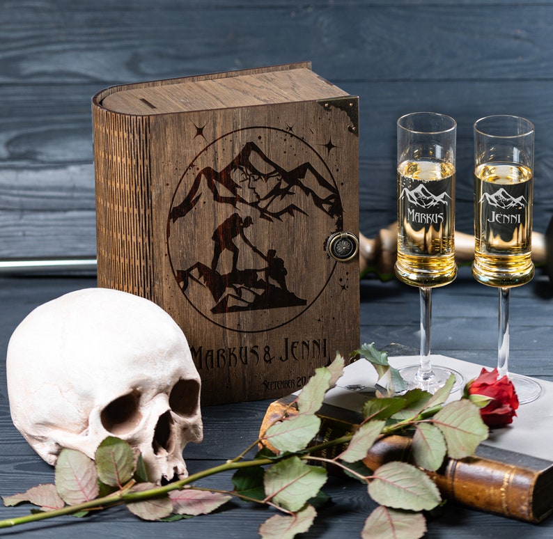 May include: A wooden box with a laser engraved design of two figures hiking in mountains. The box is closed and has a metal latch. The box is sitting on a black surface with a skull and a rose.