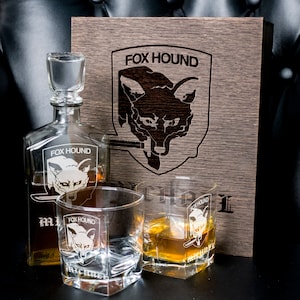 May include: A wooden box with a foxhound logo engraved on the front. The box contains a glass decanter and two glasses, all with the same foxhound logo.