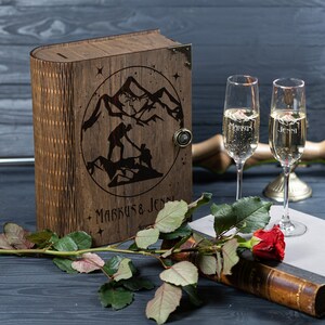 May include: A wooden box shaped like a book with a mountain scene and the names "Markus & Jenni" engraved on the front. Two champagne flutes with the same names engraved on them are in the foreground.