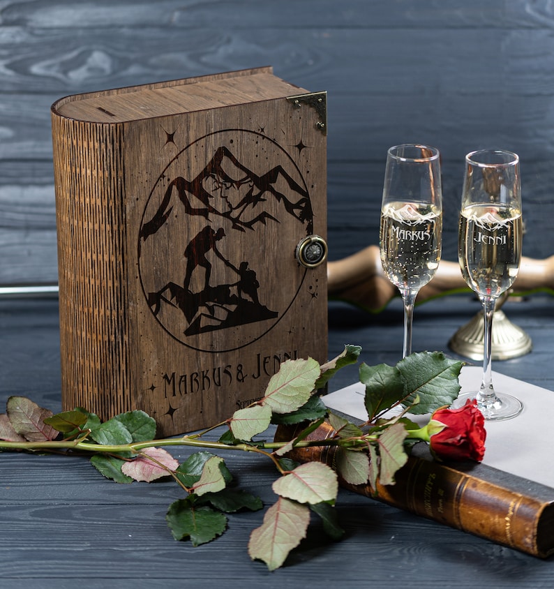 May include: A wooden box shaped like a book with a mountain scene and the names "Markus & Jenni" engraved on the front. Two champagne flutes with the same names engraved on them are in the foreground.