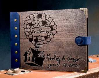 May include: A wooden photo album with a blue leather cover and brass accents. The front cover features a laser-engraved design of a house with balloons, with the text "Our Adventure" and "Mark & Jenn - April 15, 2016".