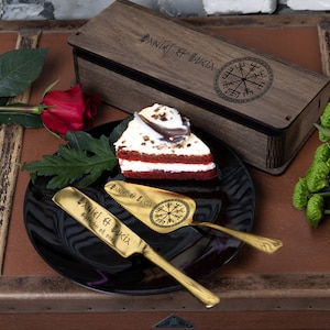 May include: A wooden box engraved with "Daniel & Lukia" and a compass design. The box contains two gold-colored cake servers with the same engraving and compass design. One server is a knife and the other is a spatula. The servers are on a black plate with a slice of red velvet cake.