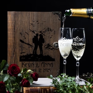 May include: A wooden wedding guest book with a mountain scene and couple silhouette is next to two champagne flutes with the names "Kevin" and "Anna". Champagne is being poured into the flutes. The date "December 12, 2025" is engraved on the book.