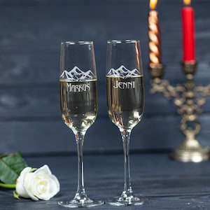 May include: Two champagne flutes with personalized engravings of "Markus" and "Jenni" with a mountain range design. The flutes are filled with champagne and are in front of a candle holder with two lit candles.
