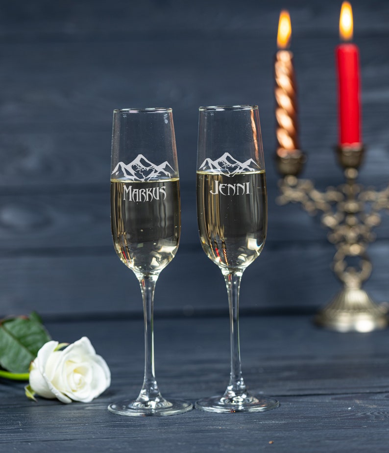 May include: Two champagne flutes with personalized engravings of "Markus" and "Jenni" with a mountain range design. The flutes are filled with champagne and are in front of a candle holder with two lit candles.