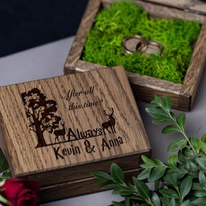 May include: Wooden ring box with a carved tree and deer design, the lid engraved with "After all this time? Always Kevin & Anna." The box is open, revealing two gold rings nestled in green moss. A romantic wedding or engagement theme.