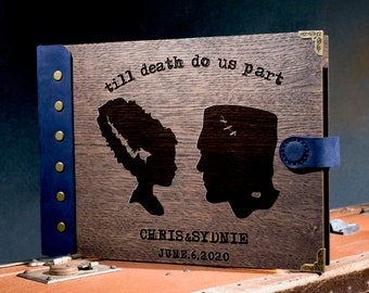 May include: A dark wood photo album with a blue leather spine and brass accents. The front cover features a laser-engraved design of a bride and groom, with the text "till death do us part" and "CHRIS & SYDNIE JUNE 6, 2020".