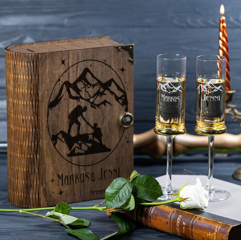 May include: A wooden box with a mountain scene and the names "Markus & Jenni" engraved on the front. Two engraved champagne flutes with the names "Markus" and "Jenni" are in front of the box.